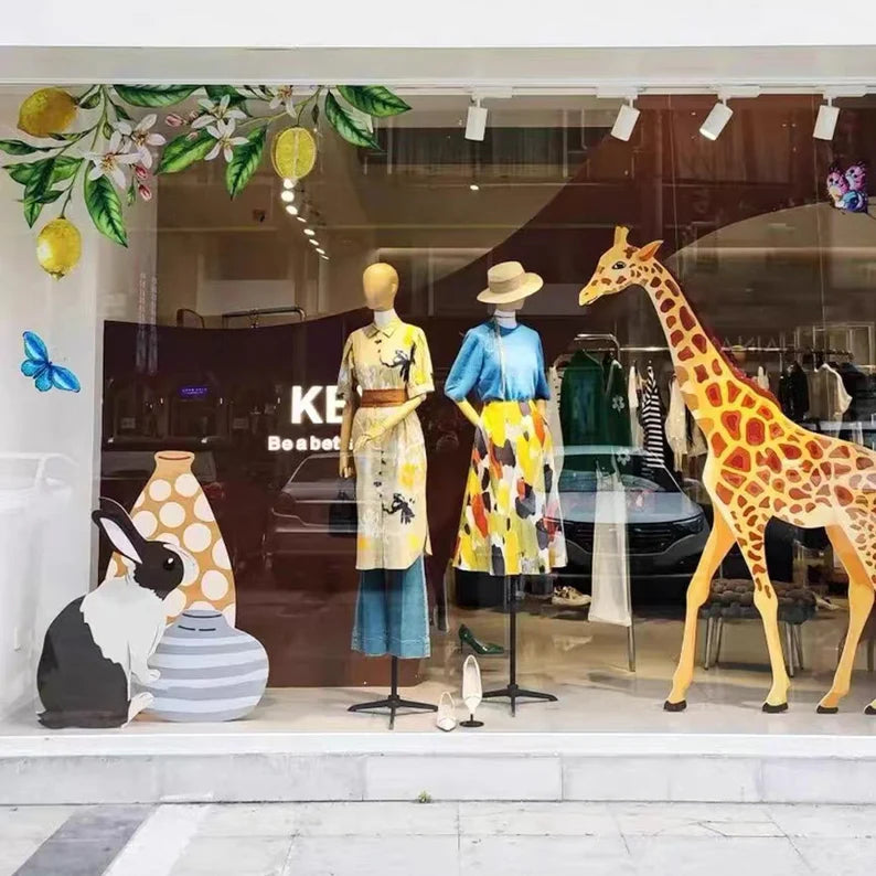 Two female display mannequins with wooden arms modeling colorful outfits in a boutique window beside a giraffe decoration.