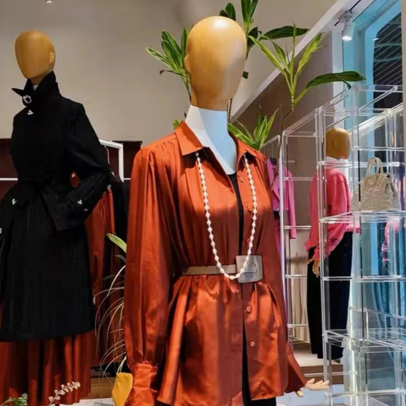 Wooden-head female display mannequin wearing a belted rust-orange blouse and long beaded necklace in a fashion boutique.